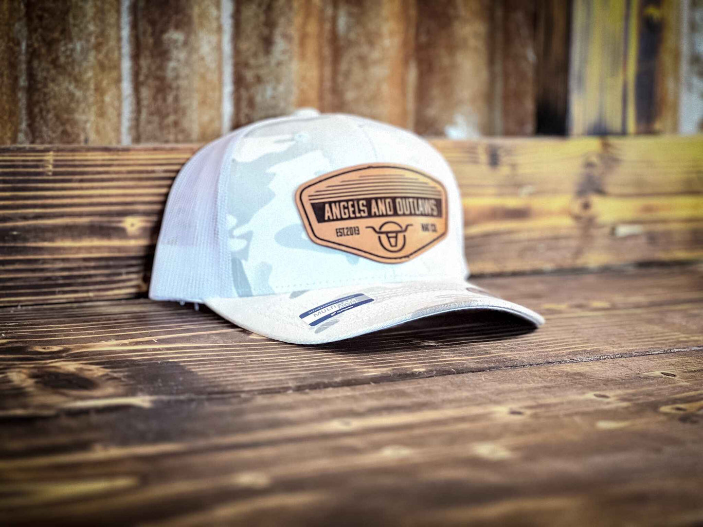 White and grey mesh back trucker hat with a leather patch that has the Angels and Outlaws Co name and log engraved on it