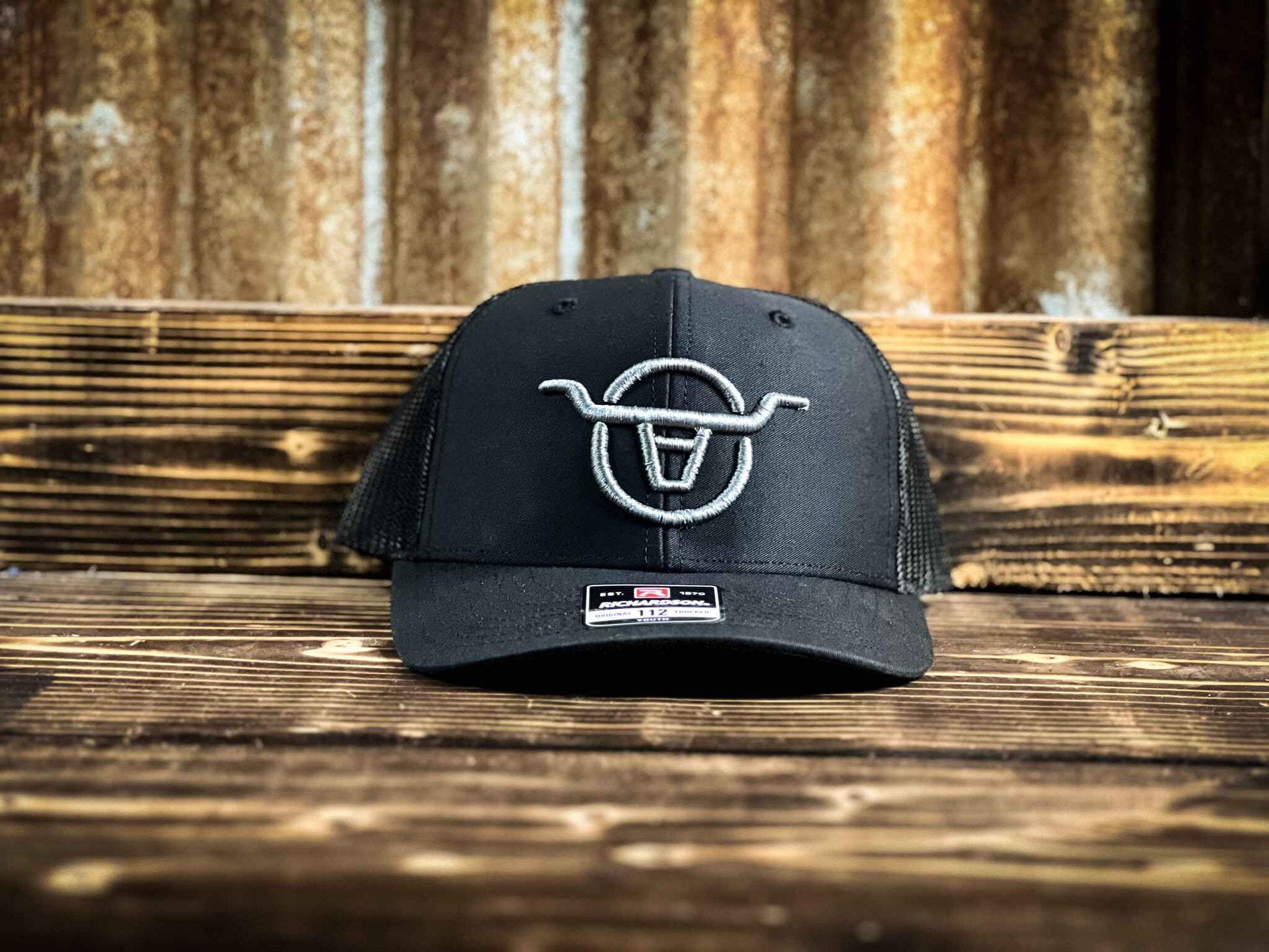 Youth size black mesh back trucker hat with Angels and Outlaws Co bull logo embroidered on the front with grey thread