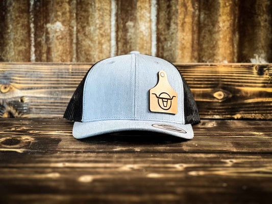 Heather grey front black mesh back trucker hat with a leather cow tag engraved with the Angels and Outlaws logo secured to hat with a rivet