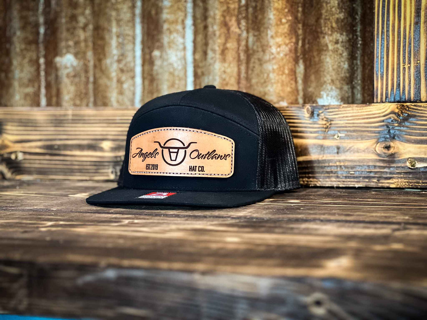 black 7 panel flat bill trucker hat with a brown leather patch with a embroidered edge that is also laser engraved with the Angels and Outlaws Co name and logo