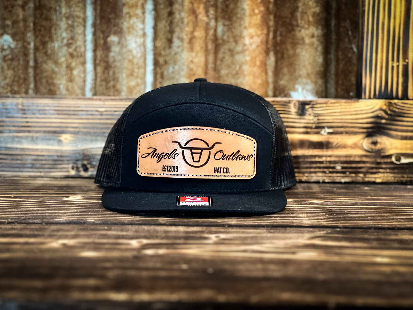 black 7 panel flat bill trucker hat with a brown leather patch with a embroidered edge that is also laser engraved with the Angels and Outlaws Co name and logo