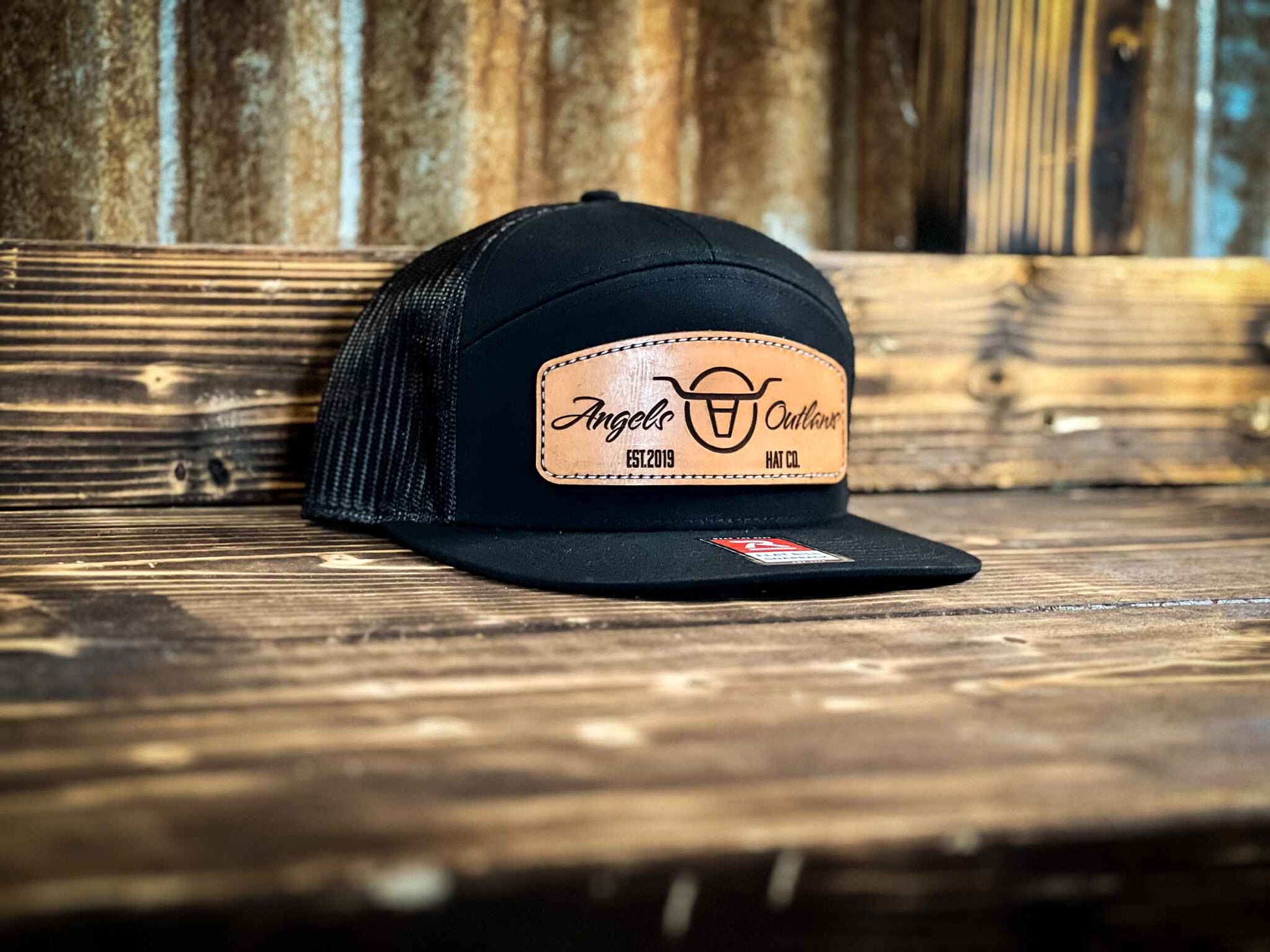 black 7 panel flat bill trucker hat with a brown leather patch with a embroidered edge that is also laser engraved with the Angels and Outlaws Co name and logo