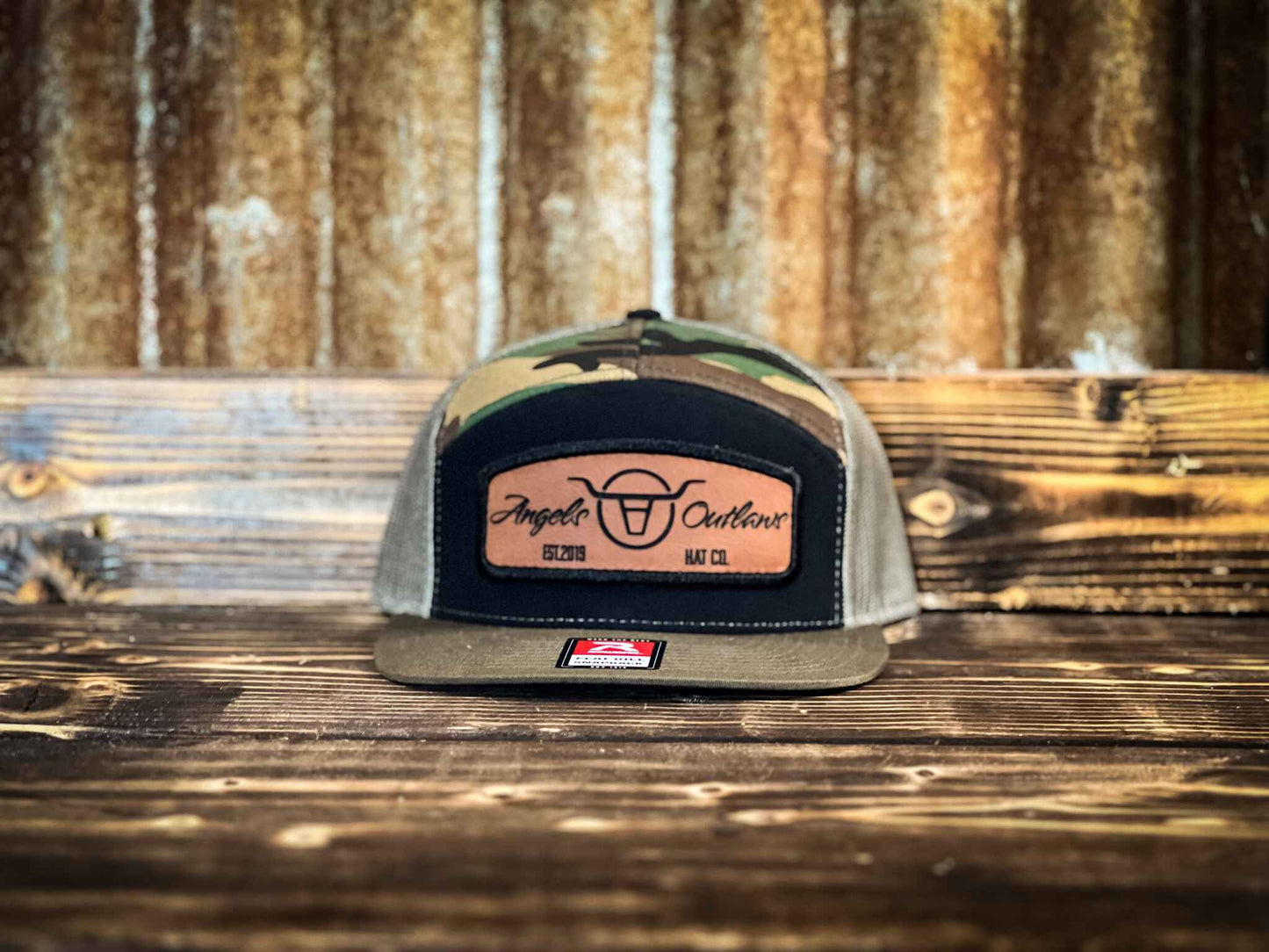 Camo 7 panel flat bill trucker hat with a brown leather patch with a embroidered edge that is also laser engraved with the Angels and Outlaws Co name and logo