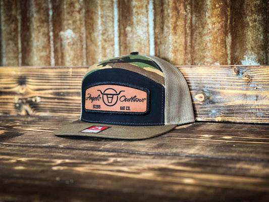 Camo 7 panel flat bill trucker hat with a brown leather patch with a embroidered edge that is also laser engraved with the Angels and Outlaws Co name and logo