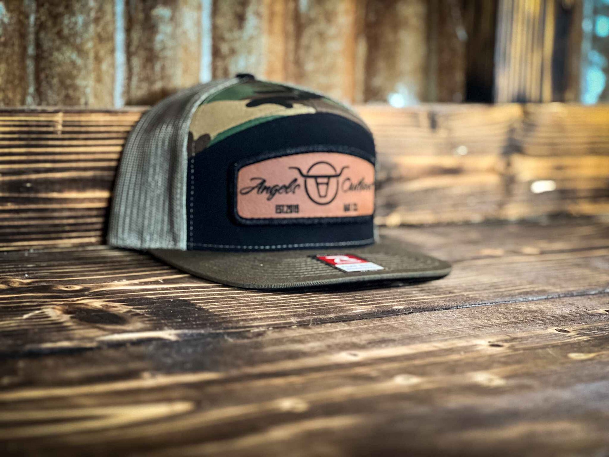 Camo 7 panel flat bill trucker hat with a brown leather patch with a embroidered edge that is also laser engraved with the Angels and Outlaws Co name and logo