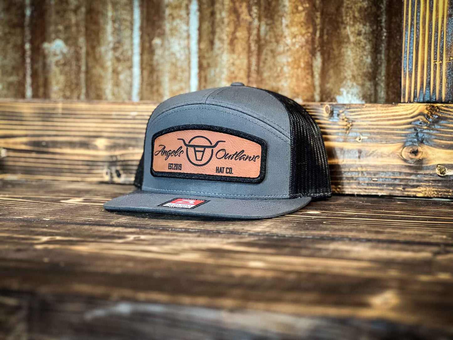 Charcoal gray front black mesh back 7 panel flat bill trucker hat with a brown leather patch with a embroidered edge that is also laser engraved with the Angels and Outlaws Co name and logo