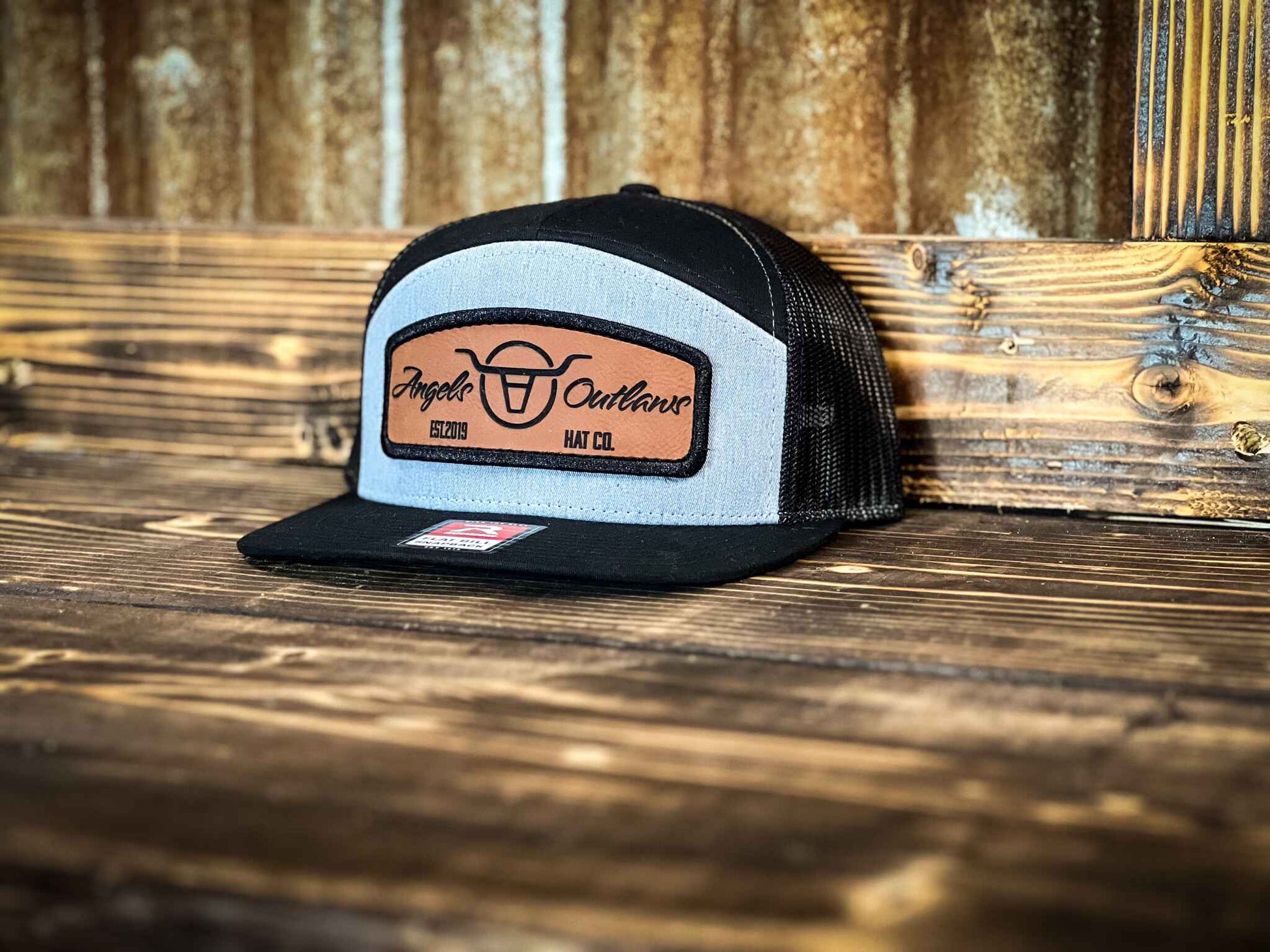 Heather gray front black mesh back 7 panel flat bill trucker hat with a brown leather patch with a embroidered edge that is also laser engraved with the Angels and Outlaws Co name and logo