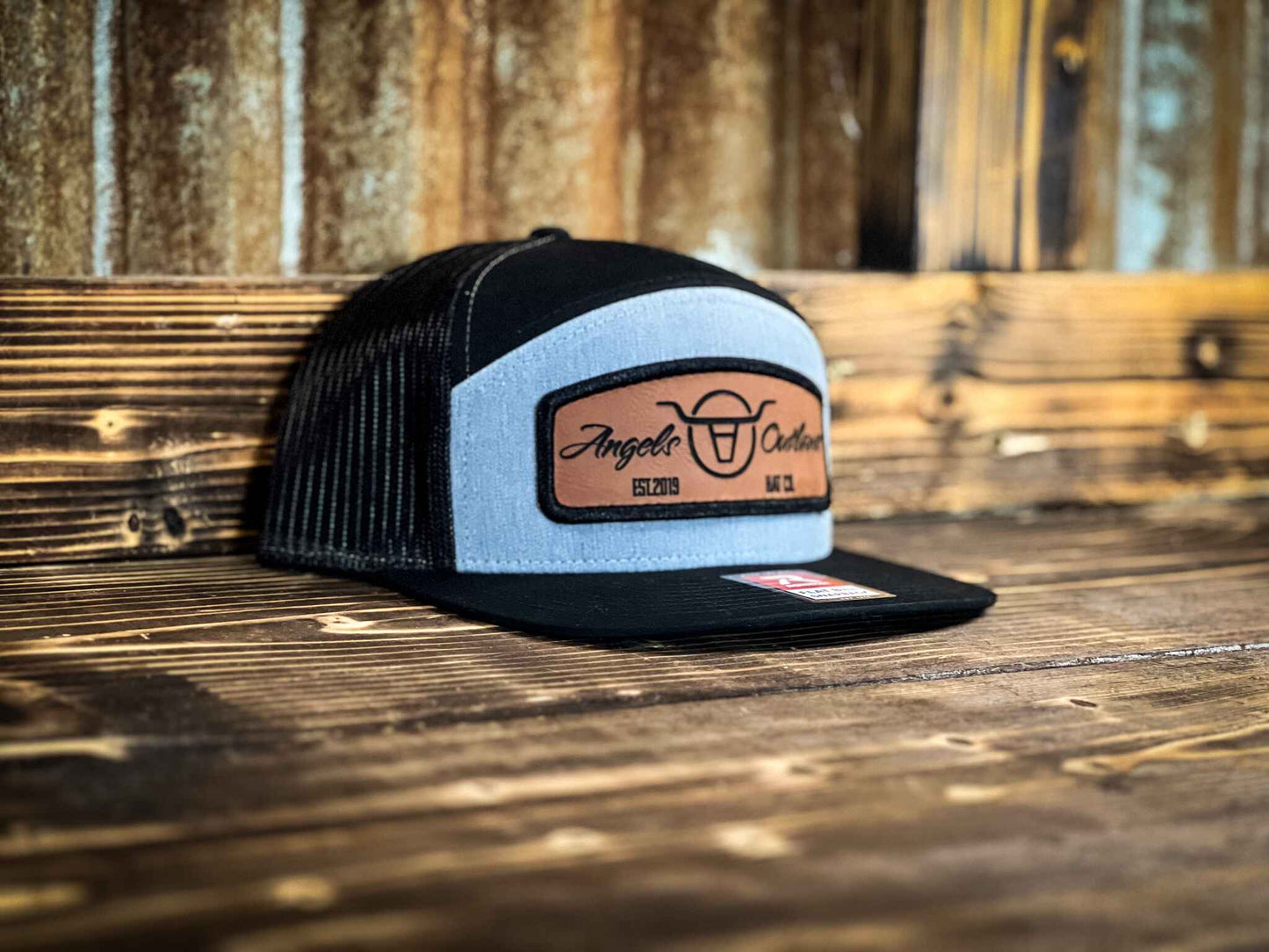 Heather gray front black mesh back 7 panel flat bill trucker hat with a brown leather patch with a embroidered edge that is also laser engraved with the Angels and Outlaws Co name and logo