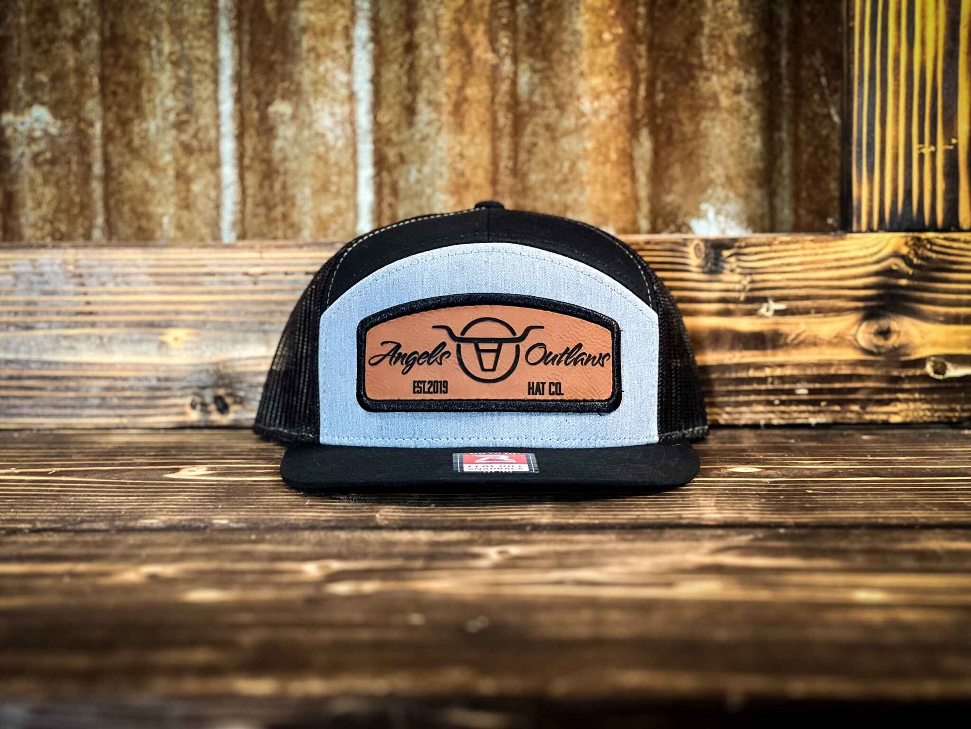 Heather gray front black mesh back 7 panel flat bill trucker hat with a brown leather patch with a embroidered edge that is also laser engraved with the Angels and Outlaws Co name and logo